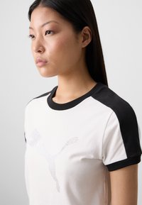 A young woman with long, straight hair poses in a black and white t-shirt featuring a rhinestone Puma logo, against a neutral background.