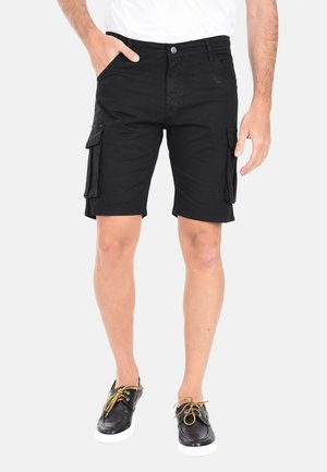 Uomo che indossa pantaloncini cargo neri e scarpe casual nere con lacci gialli, in piedi con una mano nella tasca su sfondo bianco.