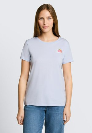 Młoda kobieta z prostymi brązowymi włosami, ubrana w jasnoniebieski t-shirt z małym kwiatowym wzorem na lewej piersi i niebieskie dżinsy.