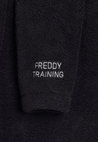 Tessuto in pile nero con una texture morbida, caratterizzato da testo ricamato bianco "FREDDY TRAINING" sul lato destro. Design semplice.