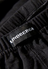 Zwarte stof met een textuur-elastische tailleband; voorzien van een geweven label met witte tekst "Lindbergh." Close-up perspectief op de details.