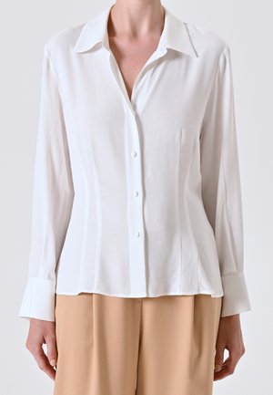 Kvinde iført en tætsiddende hvid skjorte med knapper og lange ærmer samt beige bukser med høj talje mod en ensfarvet baggrund.