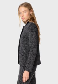 Blazer grigio scuro strutturato con scollatura rotonda, chiusura con un bottone e due tasche laterali, abbinato a una gonna coordinata.