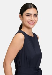 Marinblå sidoslös klänning med kroppsnära midja, sidofickor och knytbar midja. Slätt tyg med en aning lyster och minimalistisk design.