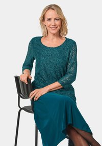 Abito teal con top in pizzo caratterizzato da un motivo testurizzato, maniche a tre quarti e una gonna fluente, abbinato a una sedia nera.