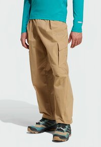 Beige cargobroek met zijzakken, een losse pasvorm en een gestikte logodetail. Draagt een blauw shirt met lange mouwen en robuuste outdoorschoenen.