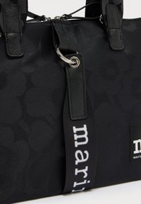 Κοντινή λήψη μαύρης πάνινης τσάντας χειρός με ανάγλυφο ανθικό μοτίβο, μαύρες δερμάτινες λαβές και μαύρο λουρί με λευκή μερική επιγραφή "marin".