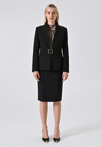 Donna in piedi con un blazer nero con cintura, gonna a tubino nera, tacchi a spillo neri con punte dorate e lunghi orecchini dorati su sfondo neutro.
