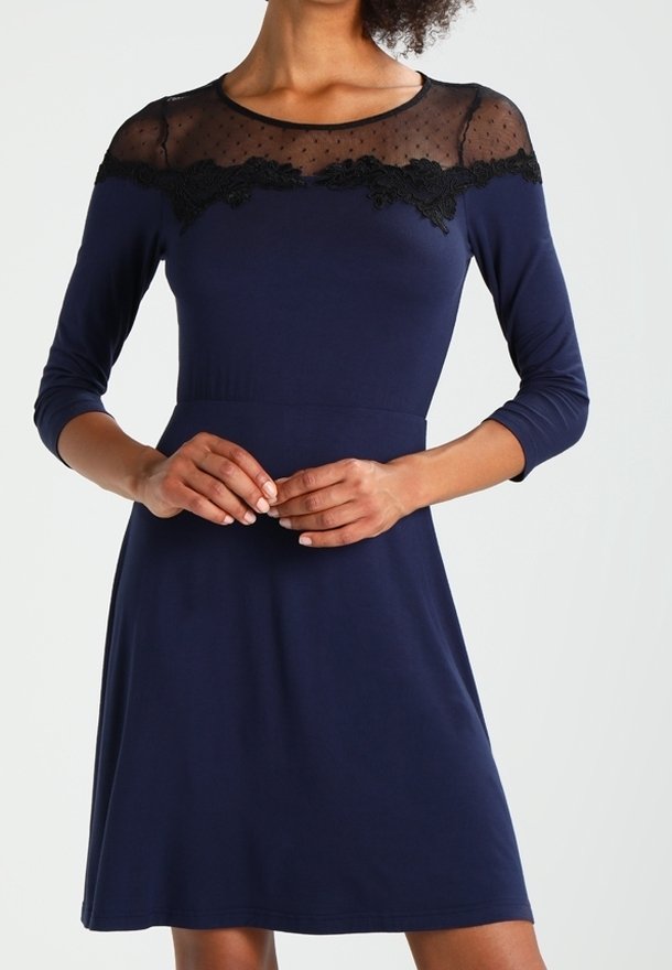 Femme portant une robe bleu marine à manches trois-quarts avec un empiècement en dentelle noire transparente sur la poitrine et les épaules.