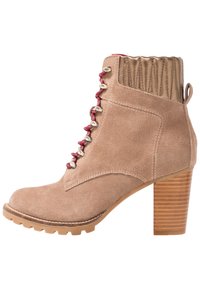 Beżowy zamszowy botek za kostkę z prążkowanym brązowym obcasem, czerwonymi sznurowadłami i metalowymi oczkami. Posiada okrągły nosek i teksturowaną tylną część.