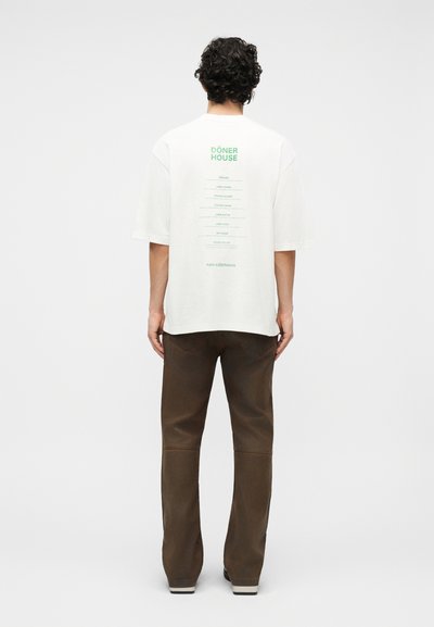 Άνδρας με σγουρά μαλλιά, φορώντας μια λευκή φαρδιά μπλούζα με πράσινο κείμενο που απαριθμεί πιάτα ντόνερ, σε συνδυασμό με καφέ παντελόνι, στέκεται σε λευκό φόντο.