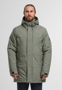 Giacca imbottita verde con colletto alto, cappuccio regolabile e due tasche frontali. Presenta dettagli in velcro nero e una patch con il logo sulla manica.