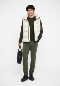 Muž v bielej vatovanej vestě přes olivovo zelený sveter a nohavice, držiaci čiernu koženú aktovku, stojaci pred svetlým pozadím.