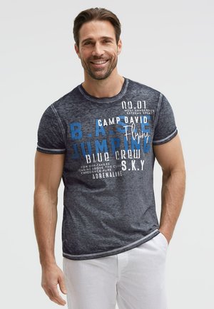 Lächelnder Mann mit kurzen dunklen Haaren, der ein dunkelblaues grafisches T-Shirt und weiße Hose trägt, steht mit einer Hand in der Tasche.