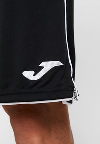 Mustat urheilushortsit, jotka on valmistettu hengittävästä kankaasta. Niissä on valkoinen logo, valkoiset sivusuunnat ja vetoketjullinen tasku.