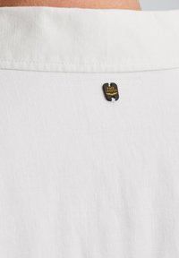 Camisa blanca con una textura a rayas sutil y una etiqueta metálica negra con el logo "PME Legend" en el cuello trasero.