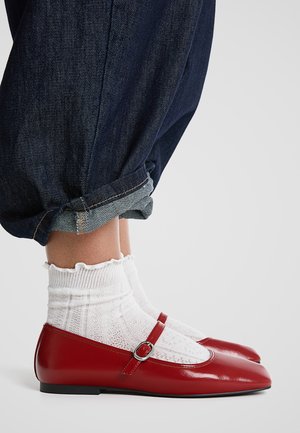 Des ballerines en cuir verni rouge avec un bout arrondi et une sangle de boucle, associées à des chaussettes blanches texturées, portées avec un jean en denim foncé retroussé.