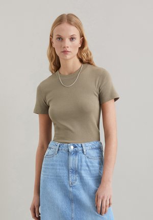 Olivegrünes geripptes T-Shirt mit kurzen Ärmeln kombiniert mit einem hellblauen Jeansrock, der einen geraden Schnitt und einen Frontknopfverschluss hat. Silberne Kettenhalskette.