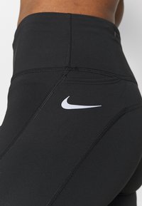 Mustat korkea vyötäröiset urheiluleggingit joustavasta kankaasta, joissa on näkyvät saumat ja valkoinen Nike-logo vasemmassa lantiossa.