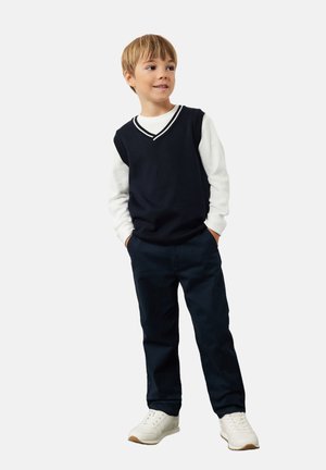 Navyblauw gebreid vest over een wit longsleeve shirt, gecombineerd met donkere broek en witte sneakers. Heeft een v-hals en contrasterende strepen.