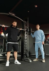 Due uomini in abbigliamento sportivo si trovano in una palestra. Uno indossa una felpa nera con cappuccio e pantaloncini; l'altro indossa un pullover azzurro chiaro e pantaloni.