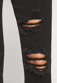 Jeans desgastados negros con dos grandes desgarros en la rodilla y bordes deshilachados, que muestran secciones de piel y capas de hilos.