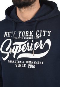Navyblauwe hoodie met witte tekst. Bevat trekkoorden met zilveren metalen uiteinden, een buidelzak en een zachte stofstructuur.
