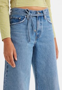 Gros plan sur une personne portant un jean bleu taille haute avec une ceinture en denim nouée et un haut court jaune à manches longues.