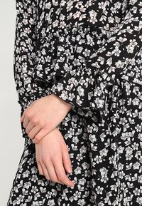 Musta kangaspaita, jossa on valkoinen kukkakuosi, varustettuna röyhelöillä hihoilla ja kerrotulla vyötäröllä, esittelee pehmeää tekstuuria ja kevyttä muotoilua.