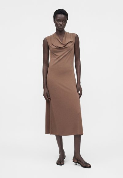 Γυναίκα φοράει ένα καφέ midi φόρεμα χωρίς μανίκια με ντραπαριστό ντεκολτέ και μαύρα σανδάλια με χαμηλό τακούνι, στέκεται μπροστά σε λευκό φόντο.