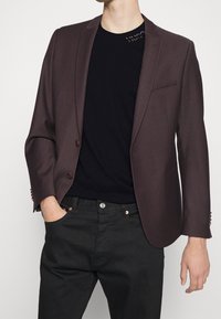 Tmavě hnědý oblekový blazer s klopami, se dvěma knoflíky a texturovanou látkou; nošený přes černé tričko a tmavé kalhoty.