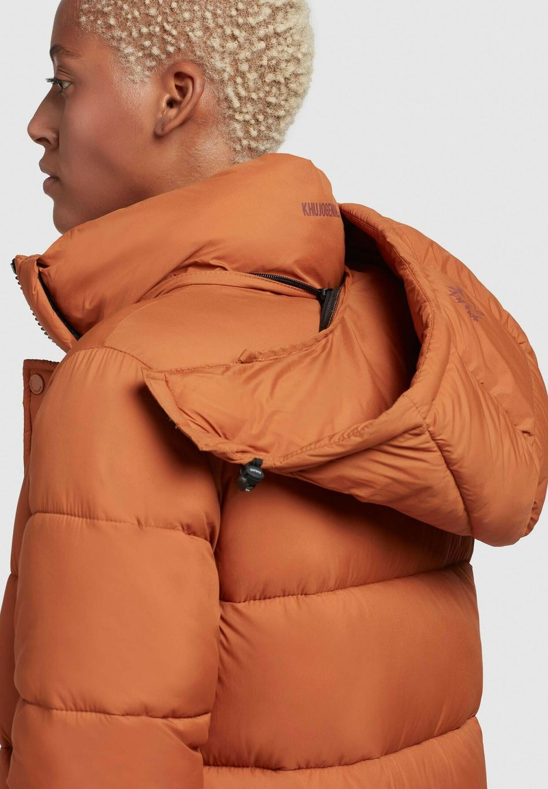 Braune Pufferjacke mit hohem Kragen, Reißverschlusstaschen und auffälligen Nähten. Strukturiertes Material mit matter Oberfläche.