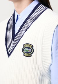 Biały dziany bezrękawnik z głębokim dekoltem w kształcie litery V, z teksturowanym wzorem granatowo-białych pasków oraz zieloną naszywką z logo Lacoste.