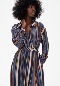 Pasiasta sukienka w granatowym, beżowym i brązowym kolorze. Posiada kołnierzyk, długie rękawy, dopasowaną talię oraz akcenty z guzikami. Gładka faktura.