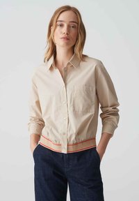 Beige knoopshirt met een borstzak en een elastische onderrand met een rode accenten. Lange mouwen met manchet details. Soepele katoenen stof.
