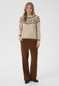 Pull en maille beige clair avec un détail de patte d'oie et un pantalon en velours côtelé marron à jambes larges. La tenue présente une coupe confortable et un design décontracté.