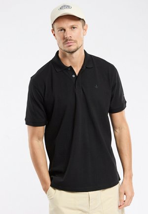 Man wearing beige cap, black short-sleeve polo shirt, and light khaki pants standing with one hand in pocket.