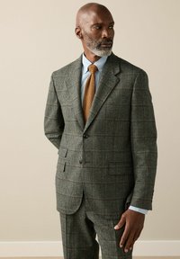 Man with gray beard wearing gray plaid suit, light blue shirt, and textured brown tie standing against beige wall, looking to side.