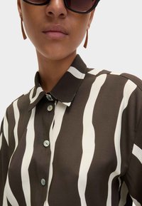 Camicia a righe marrone e crema con colletto, dotata di bottoni blu rotondi e una trama di tessuto liscia.