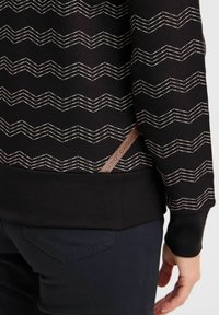 Pullover nero con un motivo a zigzag bianco, polsini e cinturino a coste. Presenta un'etichetta marrone con logo all'orlo. Tessuto morbido e texture.