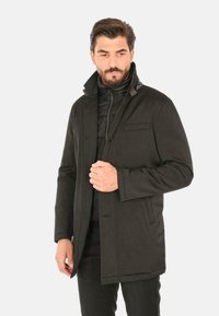 Cappotto marrone scuro con colletto, dotato di uno strato interno con zip. Il tessuto esterno è liscio, con una sola tasca sul petto e senza motivi visibili.