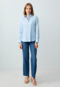 Lichtblauwe blouse met lange mouwen, gecombineerd met rechtvallende blauwe jeans. De outfit wordt aangevuld met tan hoge hakken.