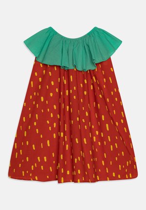 Sukienka odsłaniająca ramiona z dużą zieloną falbaną przy dekolcie, czerwoną częścią główną i rozmieszczonym pionowym wzorem w postaci żółtych kropek.