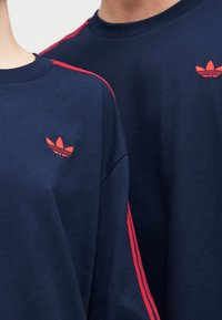 Δύο άτομα που φορούν μπλε μπλουζάκια Adidas με κόκκινες ρίγες στα μανίκια και κόκκινο λογότυπο στο στήθος, εμφανιζόμενα από το λαιμό μέχρι τη μέση του κορμού.