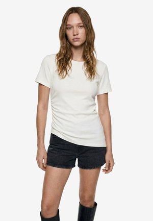 Giovane donna con capelli lunghi e mossi che indossa una maglietta bianca semplice a maniche corte, pantaloncini di jeans neri e stivali neri al ginocchio, in piedi.