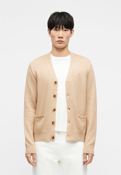 Cardigan beige a maglia con scollo a V, cinque bottoni in legno, due tasche frontali, polsini e orlo a coste, indossato sopra una t-shirt bianca e pantaloni chiari.