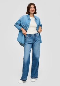 Lichtblauwe denimjacket over een witte top, gecombineerd met wijde blauwe jeans en witte sneakers. Kenmerken zijn een knoopsluiting en versleten details.