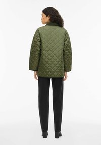 Veste matelassée vert olive avec un col, des manches larges et des coutures en forme de losange. Associée à un pantalon noir fuselé et des bottes à talons.