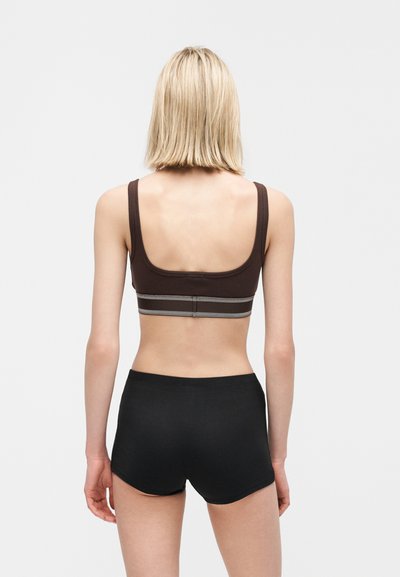 Γυναίκα με κοντά ξανθά μαλλιά που φοράει καφέ αθλητικό σουτιέν και μαύρο σορτς, εμφανιζόμενη από πίσω με φόντο μια απλή γκρι επιφάνεια.