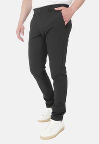 Pantaloni neri slim-fit con una texture liscia, dotati di chiusura con bottone e tasche frontali, abbinati a sneakers bianche.